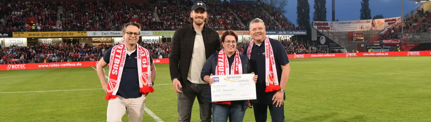 Vier Personen stehen im Stadion von Energie Cottbus auf dem Rasen und halten einen Scheck hoch