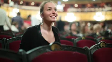 Eine junge Frau mit schwarzem Blazer sitzt in einer Stuhlreihe in einem Konzerthaus und lächelt
