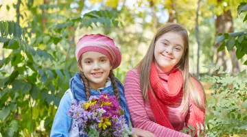 Fröhliches Mädchen mit Down-Syndrom pflückt mit Freundin Blumen auf einer Wiese im Park.