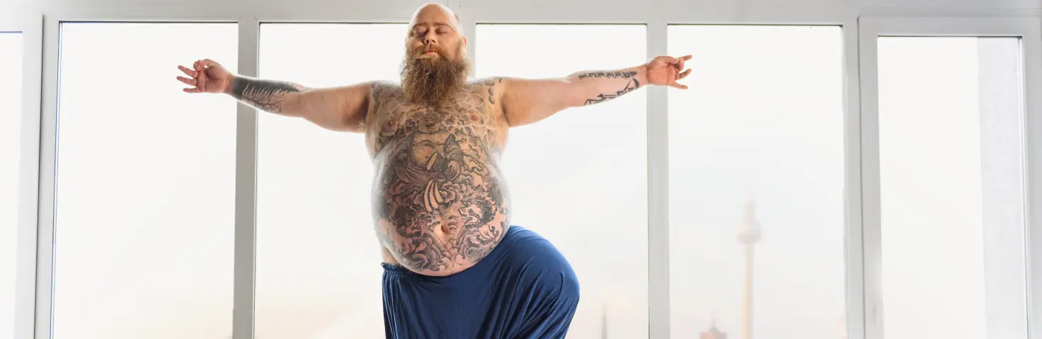 Ein beleibter Mann in blauer Stoffhose steht von einem Fenster mit Berlin-Skyline und macht Yoga.