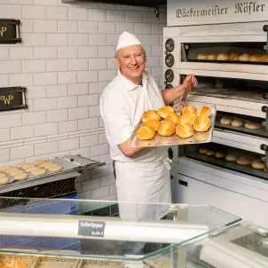 Ein Bäcker steht in einer Bäckerei und zeigt frische Brötchen.