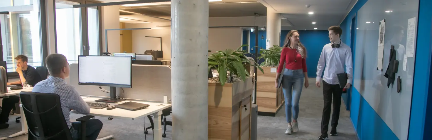 Zwei Studierende laufen im GASAG-Gebäude durch ein Großraumbüro, in dem weitere Mitarbeiter an den Schreibtischen sitzen