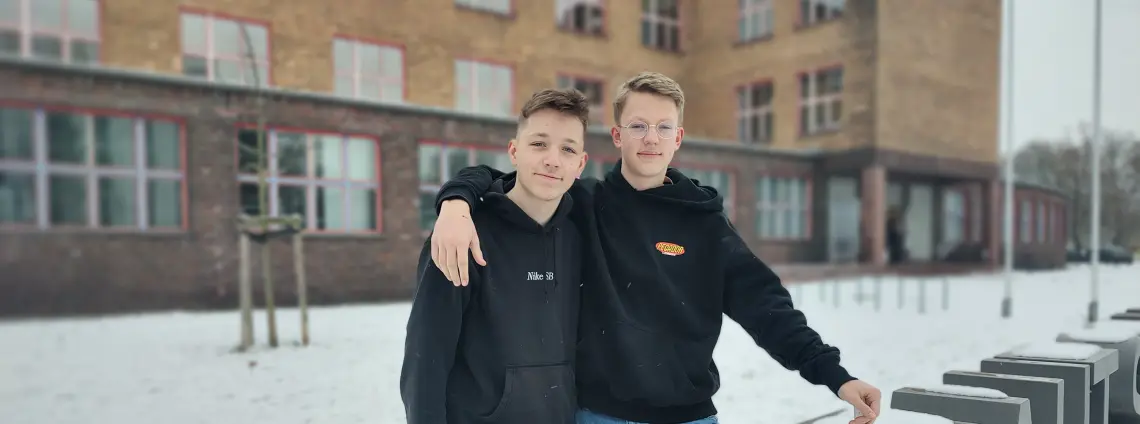 Zwei Jungs stehen vor einem Gebäude, der eine hat den Arm um den anderen gelegt. Auf dem Boden liegt Schnee.
