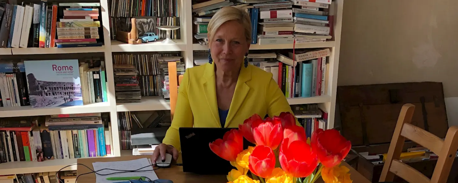 Birgit Jammes sitzt an einem Holztisch, vor ihr sind ein Laptop und ein Strauß Tulpen. Hinter ihr ist ein weißes Bücherregal.