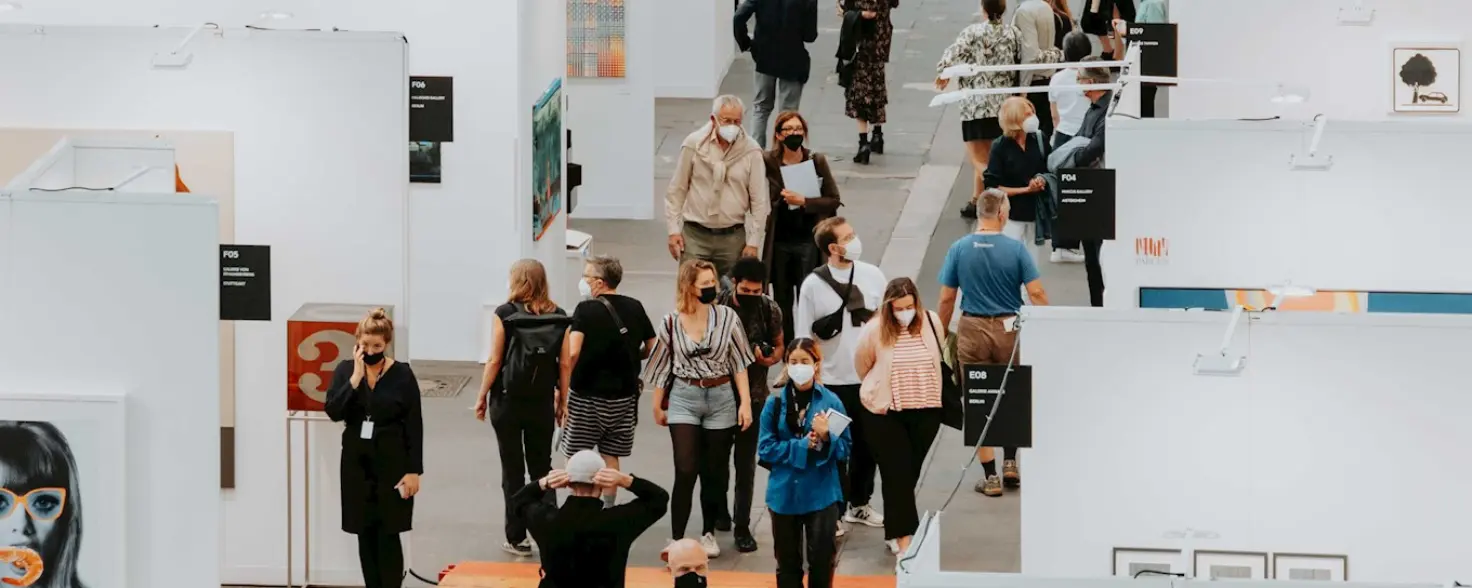 Besucher der Berlin Art Fair laufen einen Messegang entlang, links und rechts weiße Boxen, in denen Bilder ausgestellt sind.