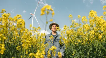 Junger Mann mit Strohhut steht in einem gelben Rapsfeld vor einer Windkraftanlage.