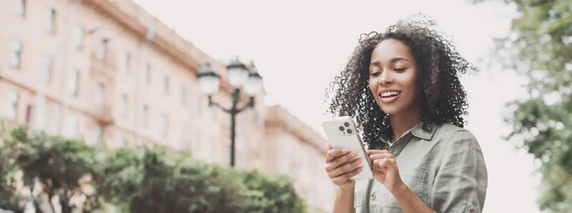 Eine Frau in städtischer Umgebung schaut lächelnd auf ihr Smartphone