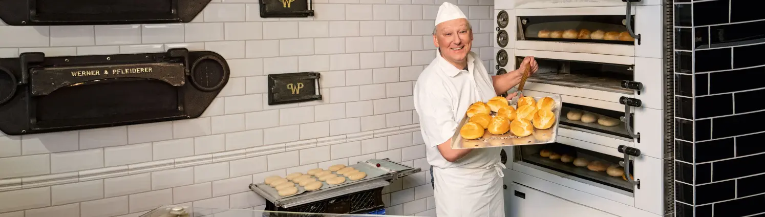 Ein Bäcker steht vor einem Ofen und präsentiert eine Bäcker-Schaufel voll frischer Brötchen